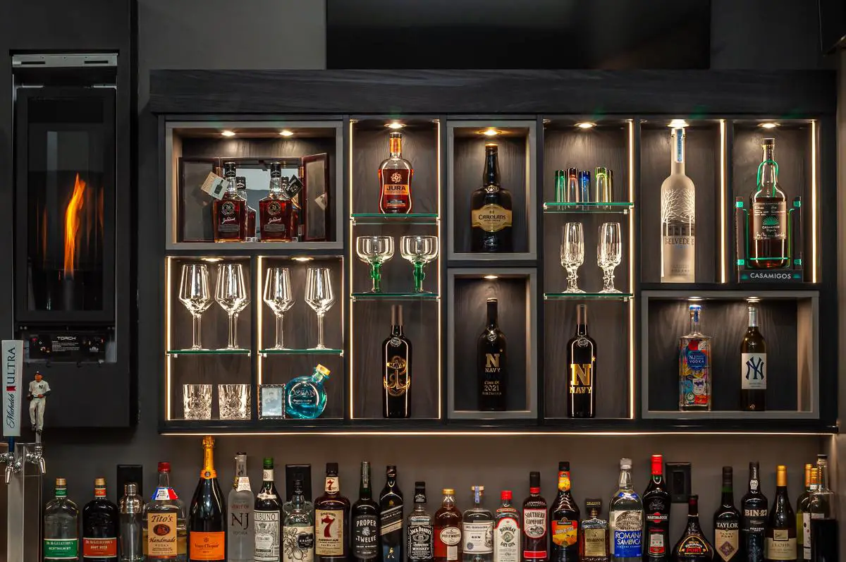 An image of a stylish home bar made with IKEA furniture, displaying shelves with bottles of alcohol and glasses, decorative coasters, and a small indoor plant.